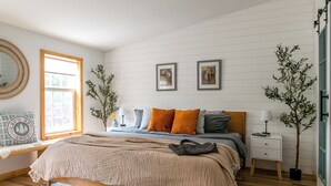 Un dormitorio con cama, mesitas de noche, una ventana y cuadros en la pared.