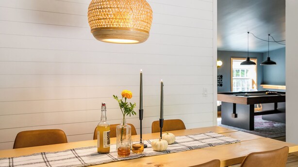 Un comedor con una mesa de madera, una lámpara colgante de fibras, velas, una botella de vino y un jarrón con flores.