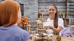 A person serving food at a wooden table with a tiered serving tray, a glass of water, and a vase with orange flowers. - Place to relax  (Zermatt)