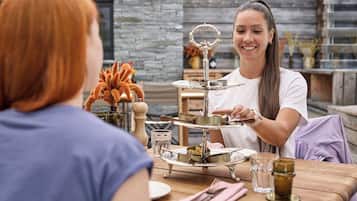 A person serving food at a wooden table with a tiered serving tray, a glass of water, and a vase with orange flowers.