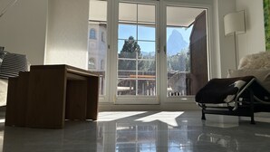 A modern living room with a large sliding glass door leading to a balcony, a wooden side table, a black leather chair, and a white lamp. - Place to relax  (Zermatt)