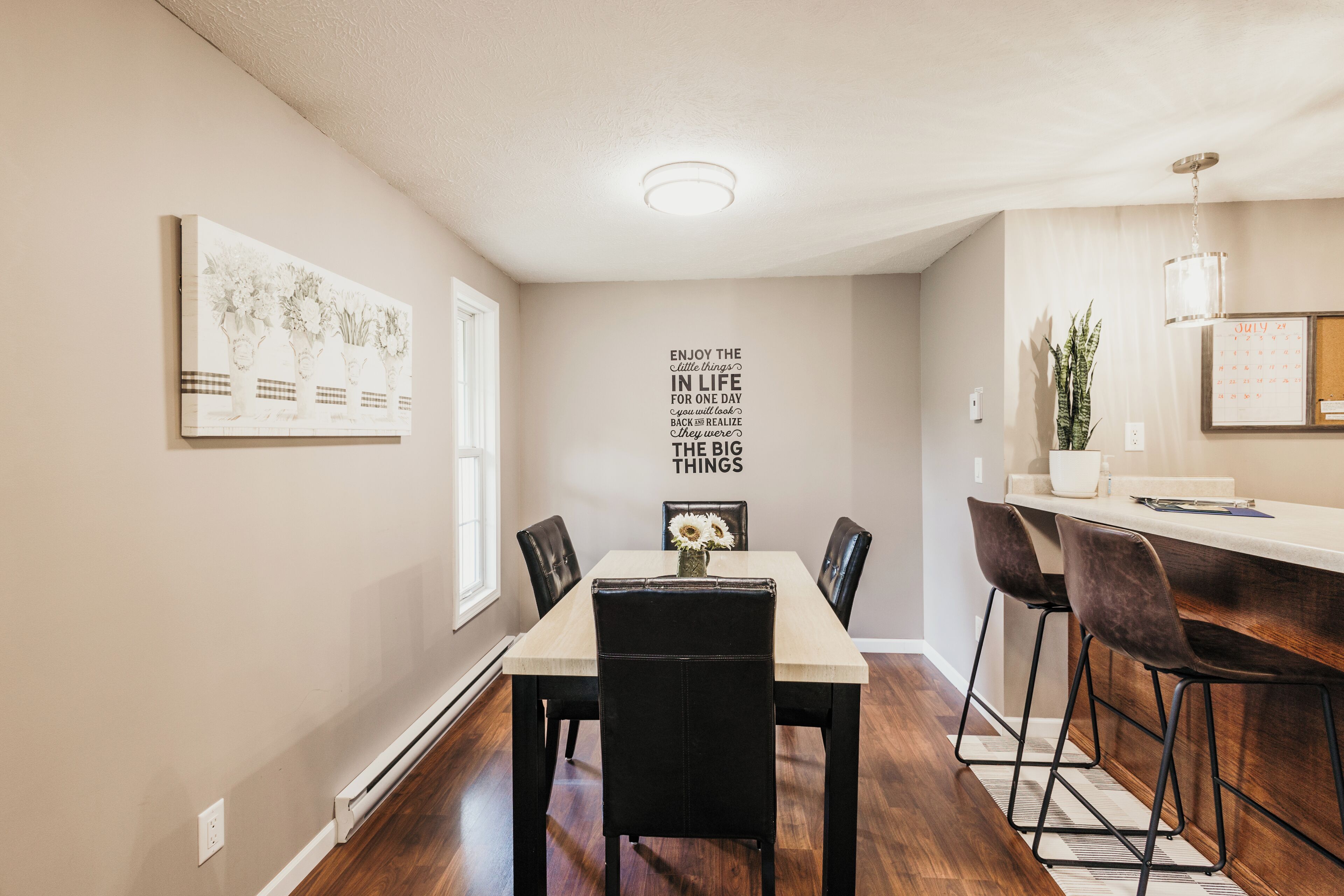 A dining area with a table, chairs, a wall-mounted calendar, and a decorative wall piece.