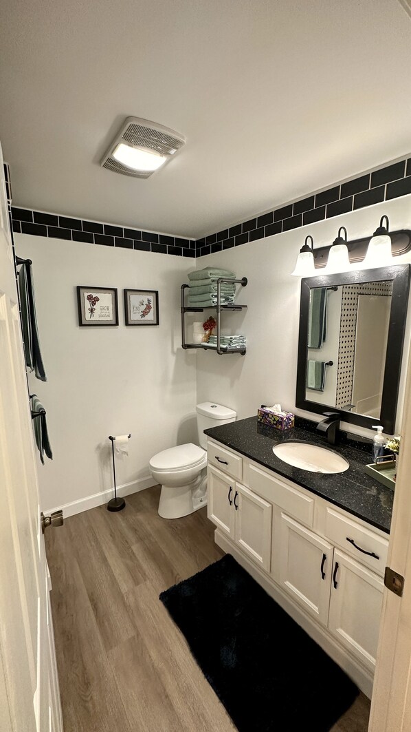 Baño con inodoro, lavabo y tocador, con zócalos blanco y negro, alfombra negra y rejilla de ventilación en el techo.