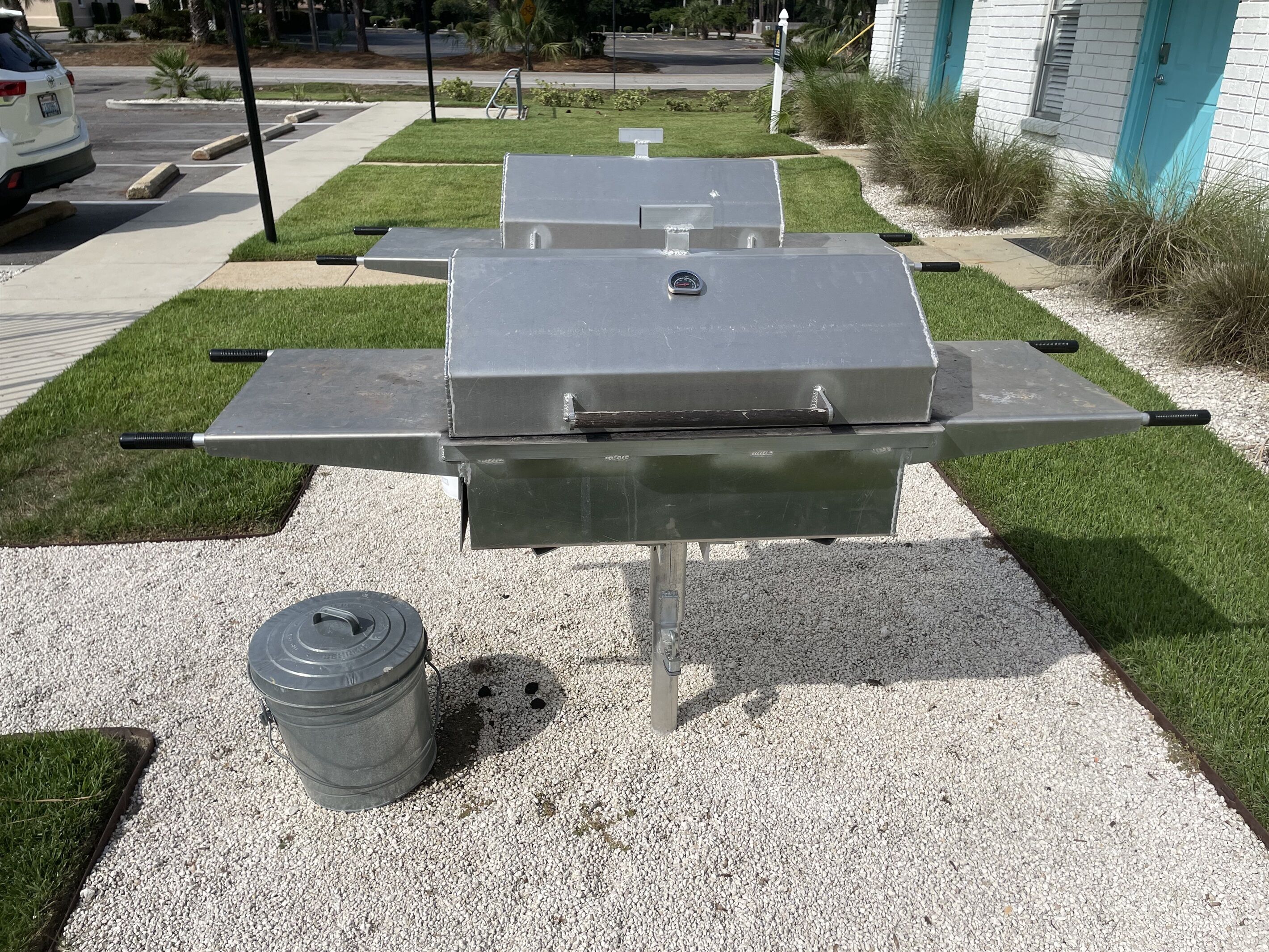 A portable grill with a lid, a handle, and a metal frame, placed on a gravel surface with grass patches around.