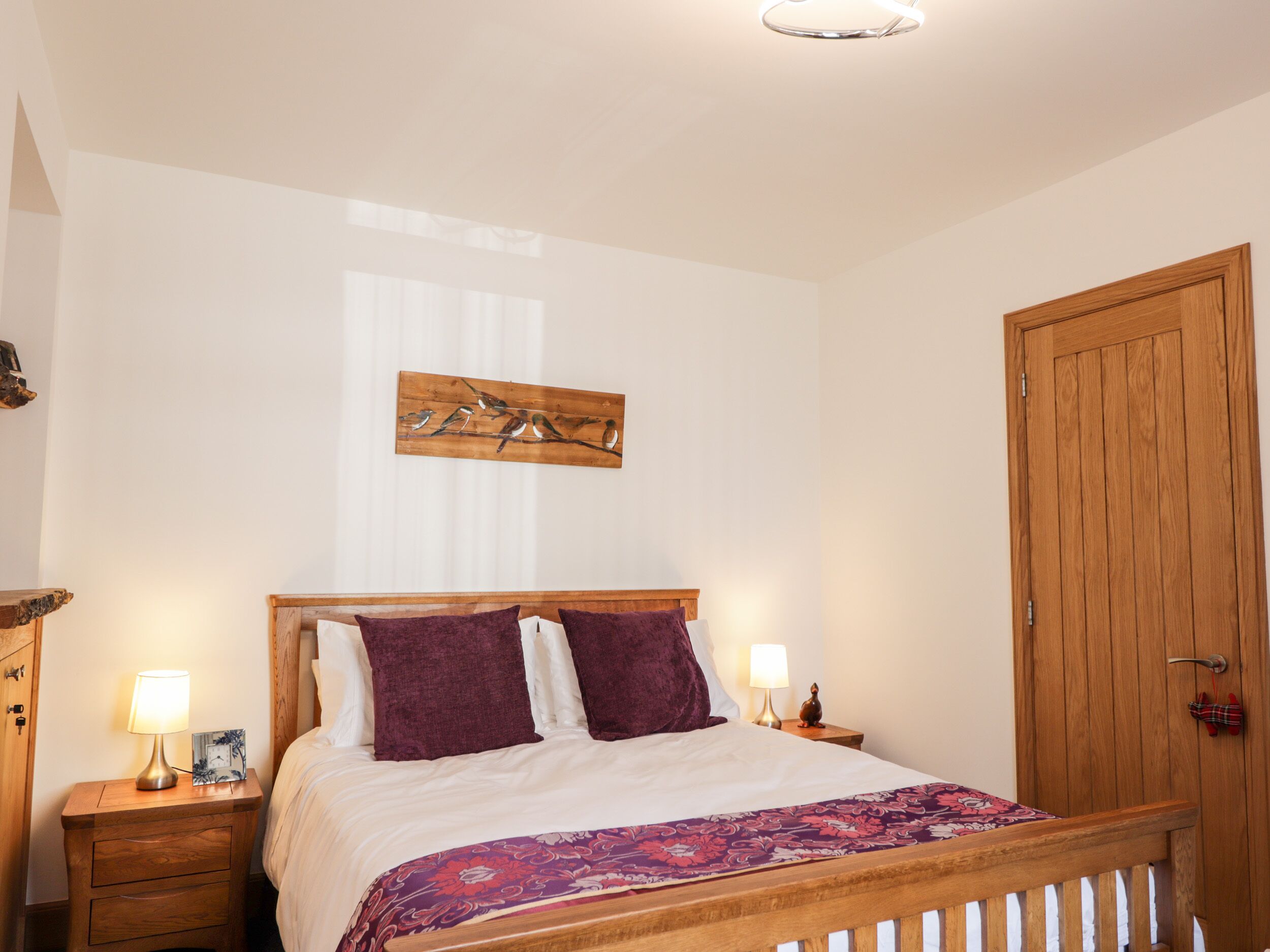 A bedroom with a wooden bed, two bedside tables with lamps, a wooden door, and a wall-mounted artwork of birds on a branch.