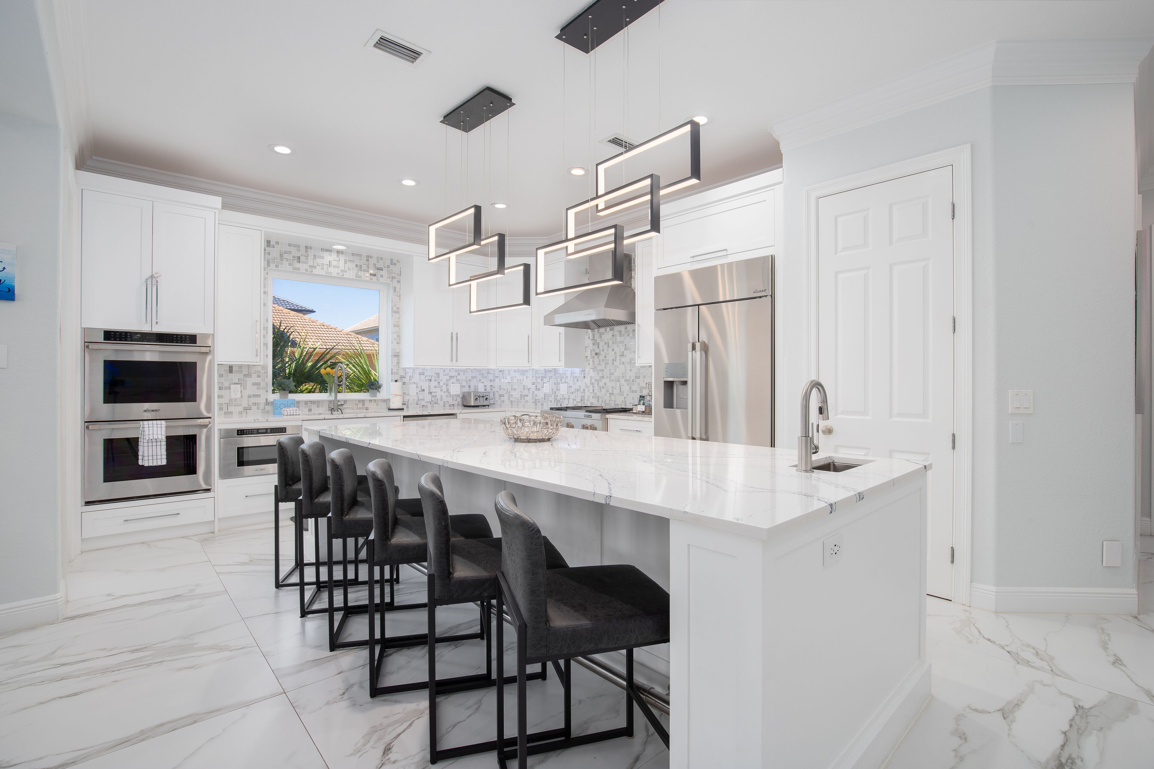 A modern kitchen with white cabinetry, a large island with bar stools, stainless steel appliances, and a view to the outdoors.