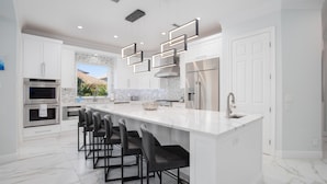 A modern kitchen with white cabinetry, a large island with bar stools, stainless steel appliances, and a view to the outdoors.