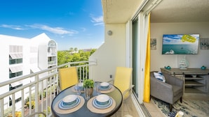 A balcony with a table set for two, chairs, a small table with decorative items, and a view of buildings and greenery. - **THE OSPREY'S NEST** Condo & Pool + Last Key White Glove Service... (Key West)