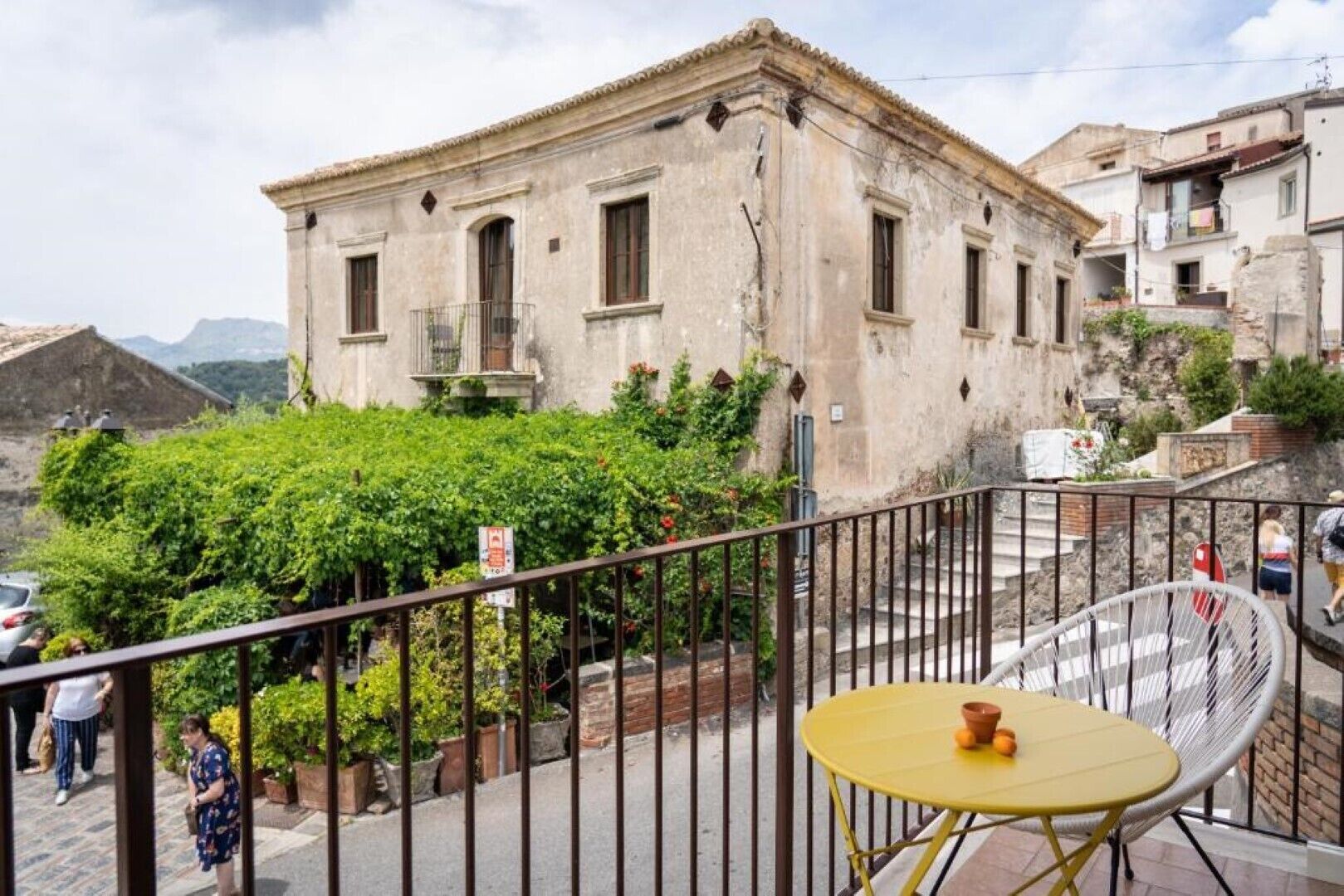 A balcony with a yellow table and a white chair, overlooking a building with a garden and a mountain in the background.