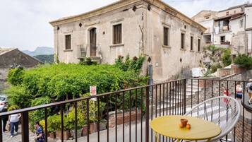 A balcony with a yellow table and a white chair, overlooking a building with a garden and a mountain in the background.
