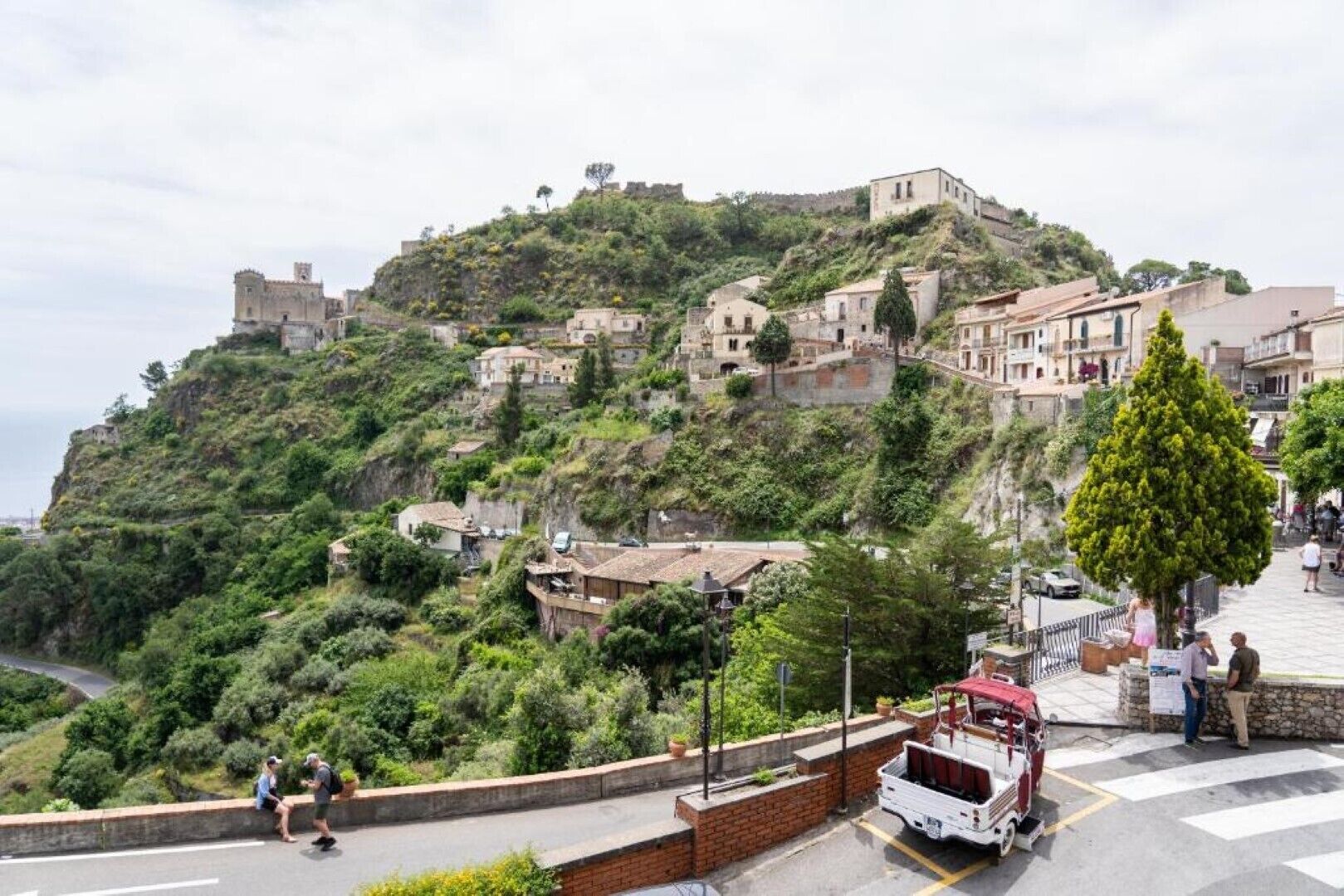 Un poble situat en un turó amb edificis històrics, un camí pavimentat i un vehicle estacionat.
