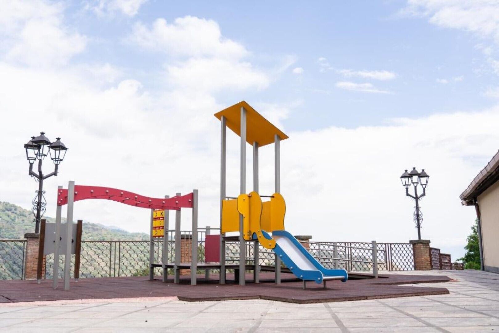 Ein Spielplatz mit Rutsche und Klettergerüst, umgeben von einem Zaun und Straßenlaternen.