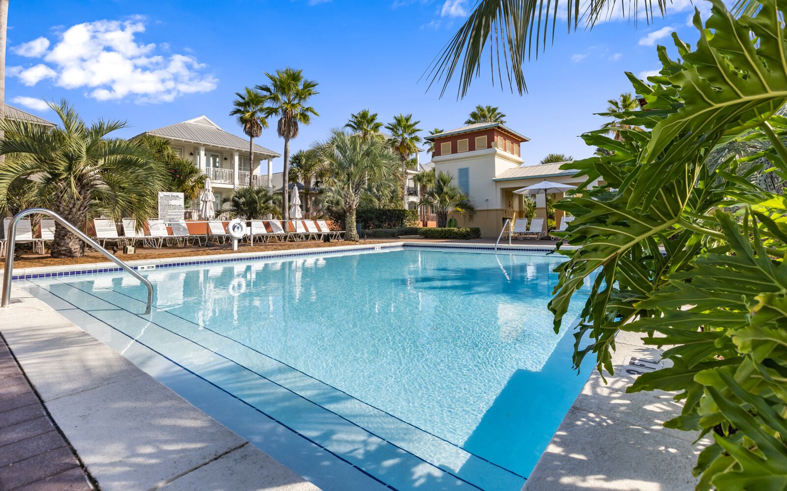 A clear blue pool surrounded by lounge chairs and palm trees, with a building in the background.