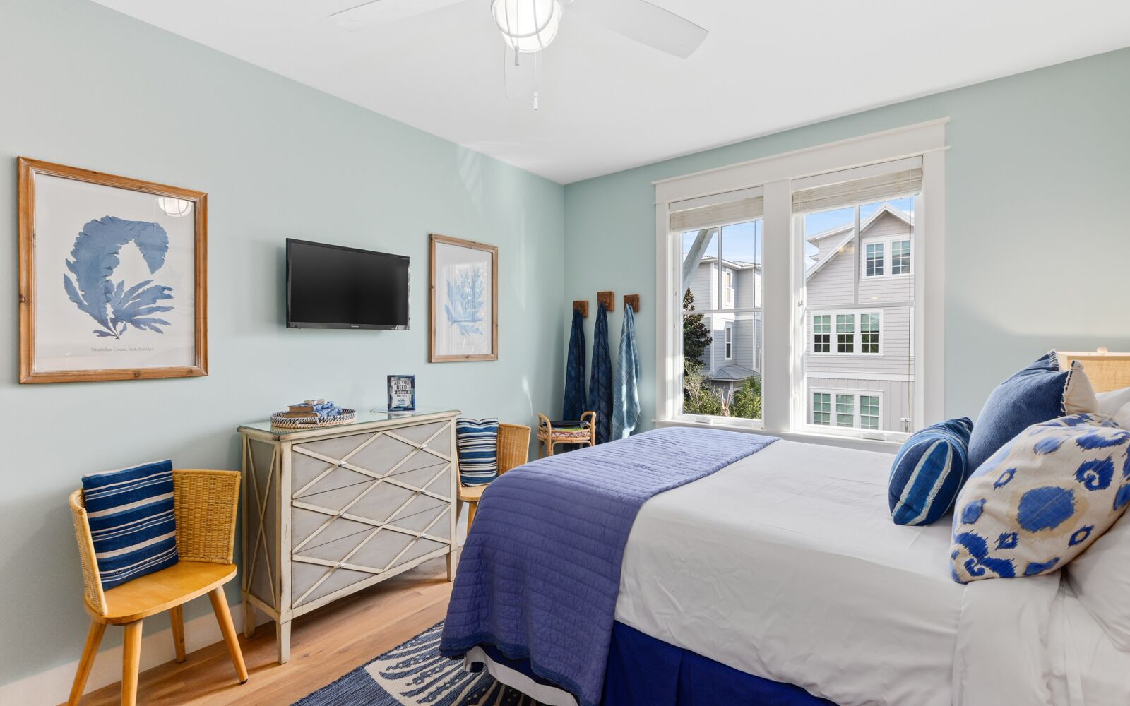 A bedroom with a bed, a dresser, a television, and a window with a view of houses.