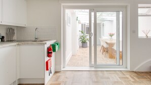 A modern kitchen with white cabinets, a sink, and a fire extinguisher. There is a dining area with a table and chairs visible through a glass door. - Cosy Gem in Covent Garden (London)