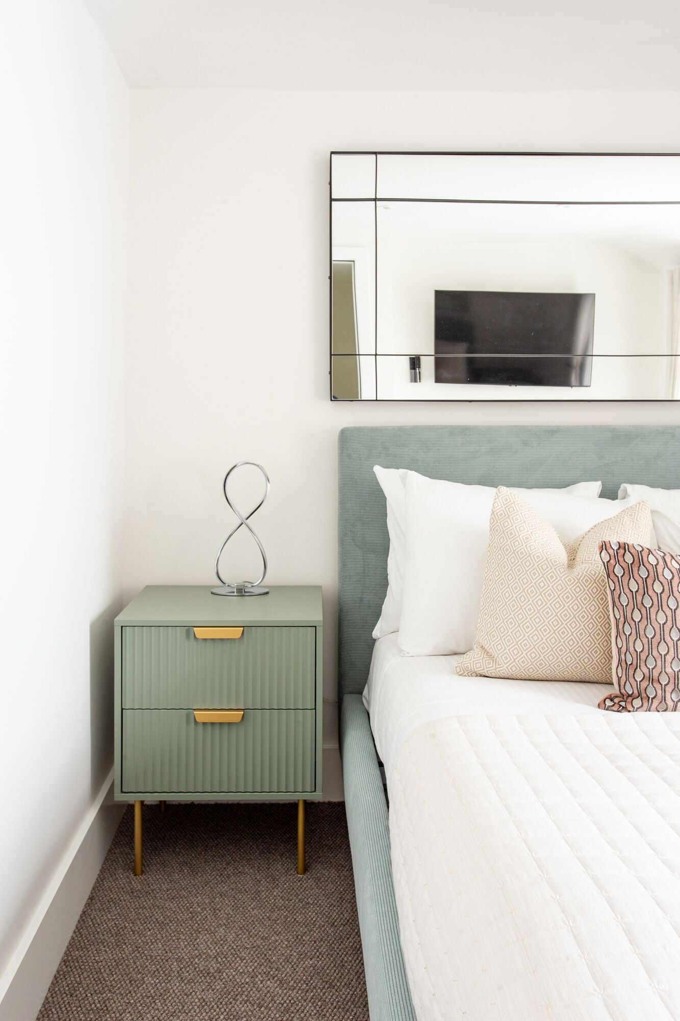 Une chambre à coucher bien rangée, avec une table de chevet verte, un lit agrémenté d’oreillers à motifs et un grand miroir placé au-dessus du lit.