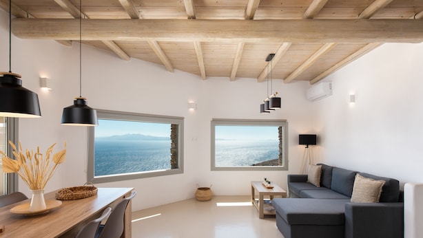 A modern living room with a wooden ceiling, large windows with ocean views, a dark grey sofa, and a wooden dining table with chairs. - Caval Residences (Tinos)