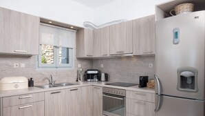 A modern kitchen with light wood cabinets, stainless steel appliances, a built-in oven, and a refrigerator with a water dispenser. - Caval Residences (Tinos)