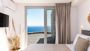 A bedroom with a bed, a TV, a potted plant, and a view of the sea through a sliding door. - Caval Residences (Tinos)