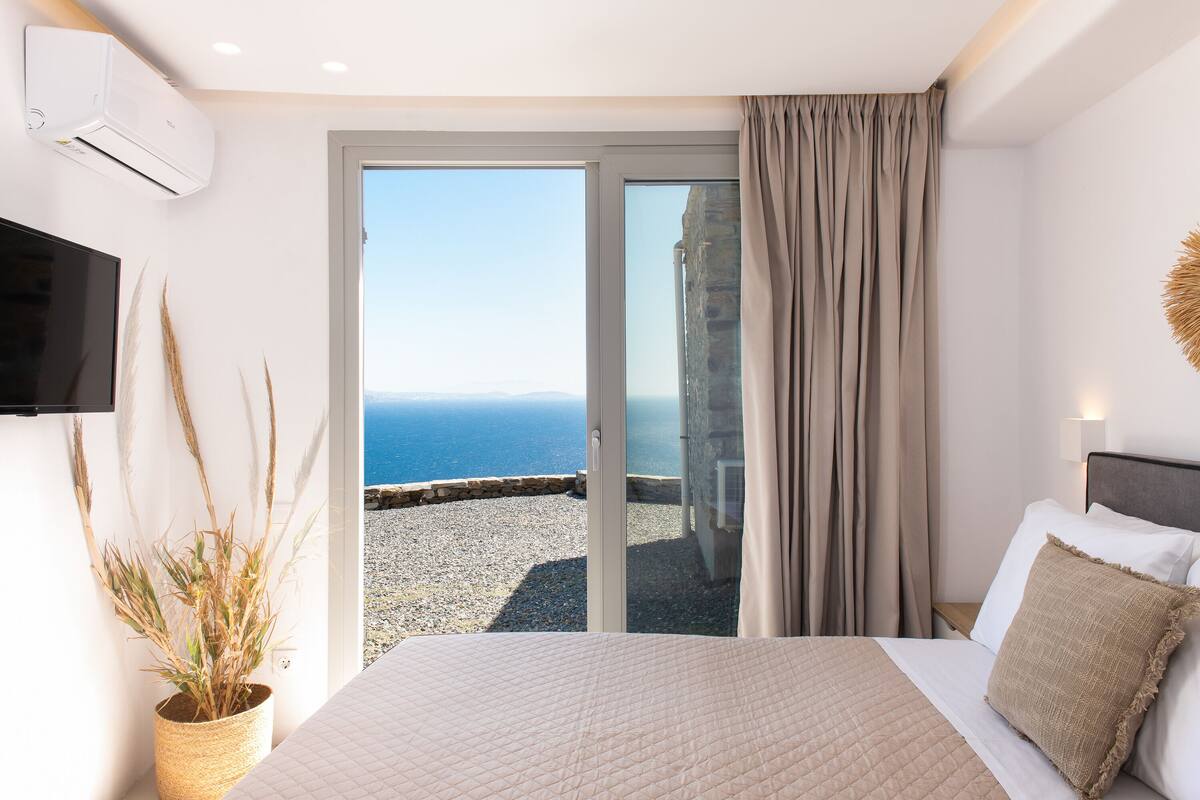 Un dormitorio con cama, televisor, planta en maceta y vista al mar a través de una puerta corrediza.