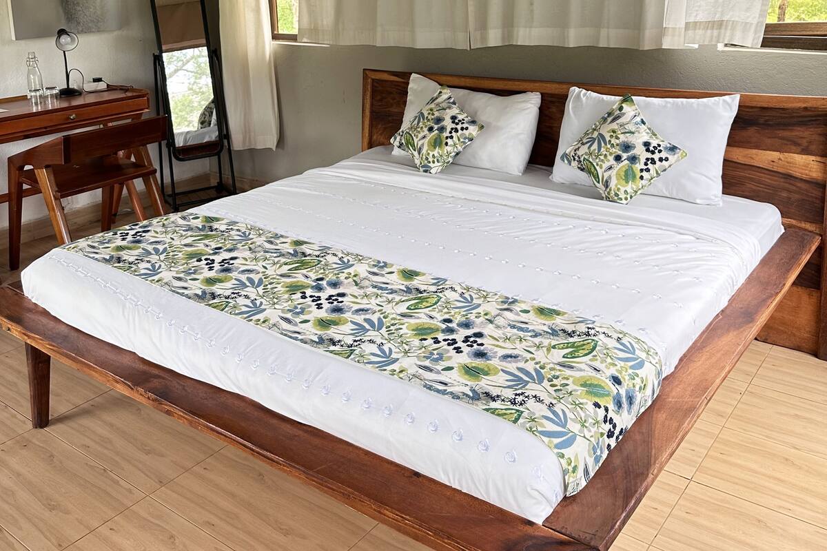 A bedroom with a wooden bed, white bedding, a desk with a chair, a mirror, and a window with curtains.