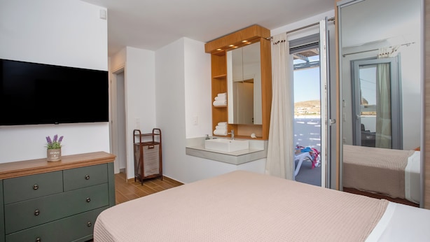 A bedroom with a bed, a TV, a dresser, a mirror, a bathroom with a sink, and a sliding door to a balcony. - Whisper Villa Mykonos (Mykonos)