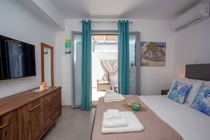 A hotel room with a bed, a wooden dresser, a TV, and a view through an open door. - Whisper Villa Mykonos (Mykonos)