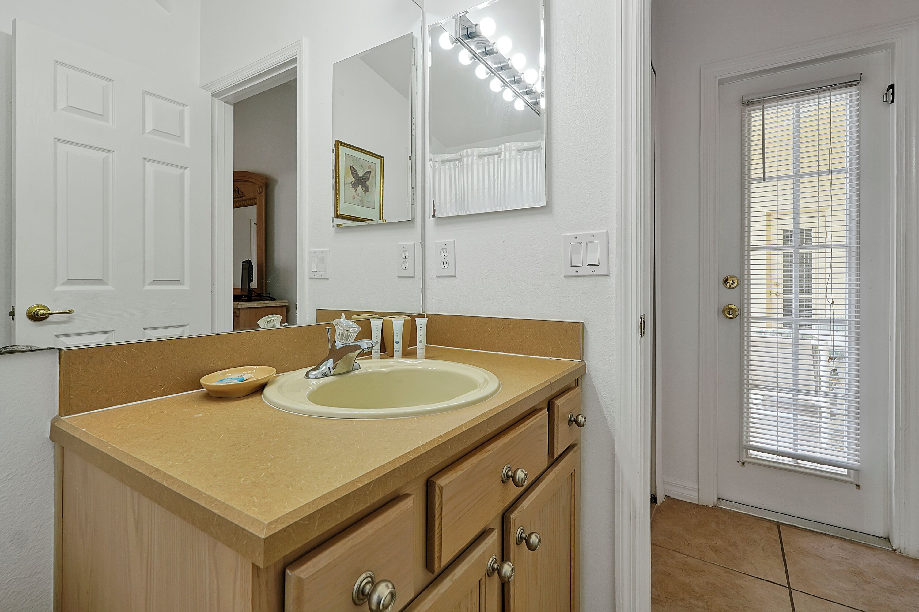 A bathroom with a beige countertop, a sink, and a mirror with lights. There is a door with a window and blinds.