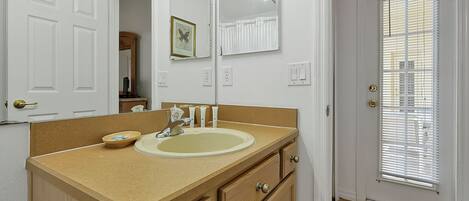 A bathroom with a beige countertop, a sink, and a mirror with lights. There is a door with a window and blinds.