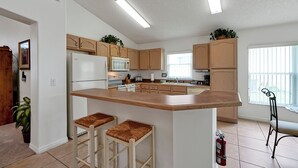 A modern kitchen with wooden cabinets, a central island with stools, a refrigerator, and a window providing natural light. - Affordable 5BR Disney Villa with Pool, Spa & Game Room - Indian Creek Community (Kissimmee)