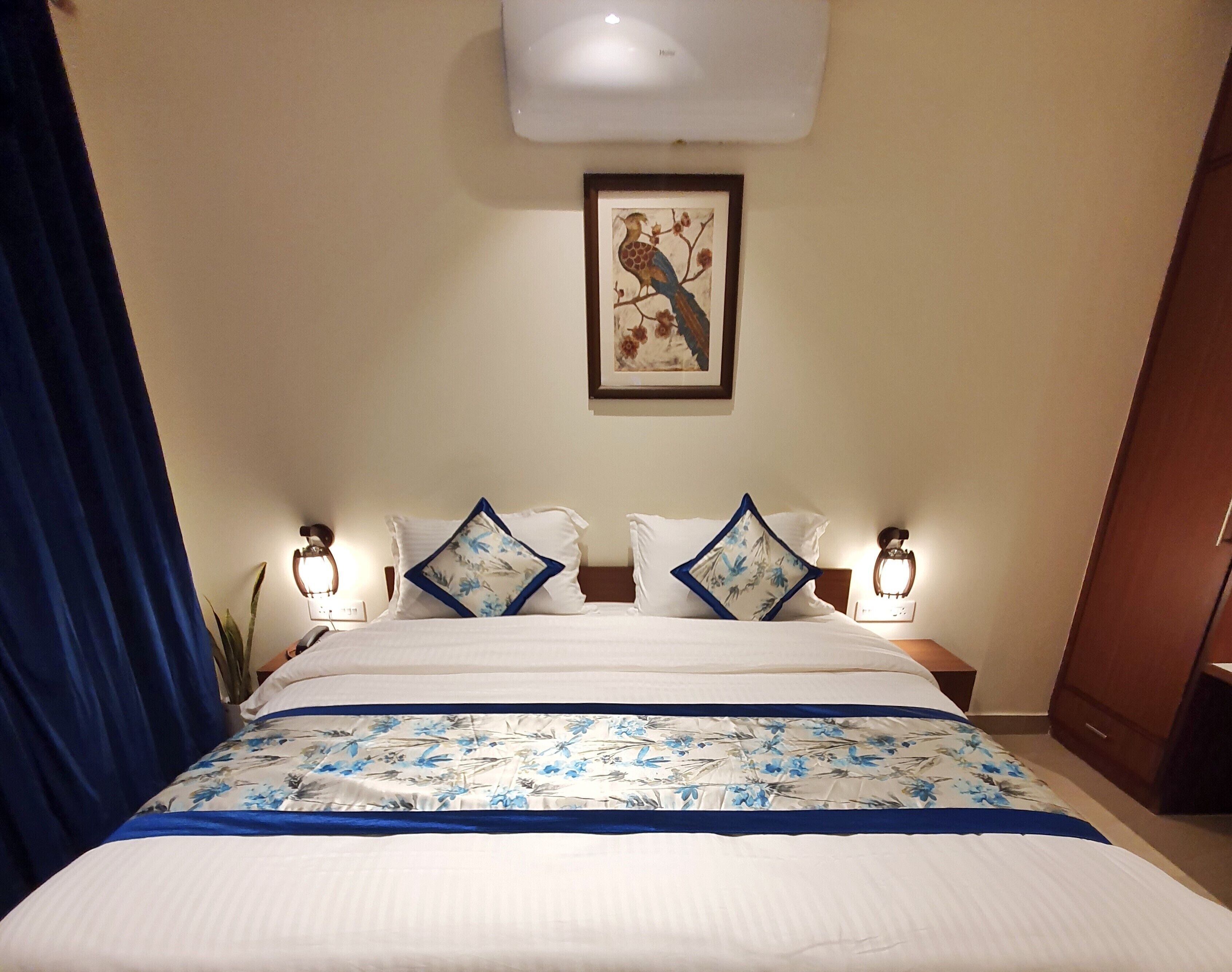 A hotel room with a large bed, two bedside lamps, a framed artwork on the wall, and a blue curtain.