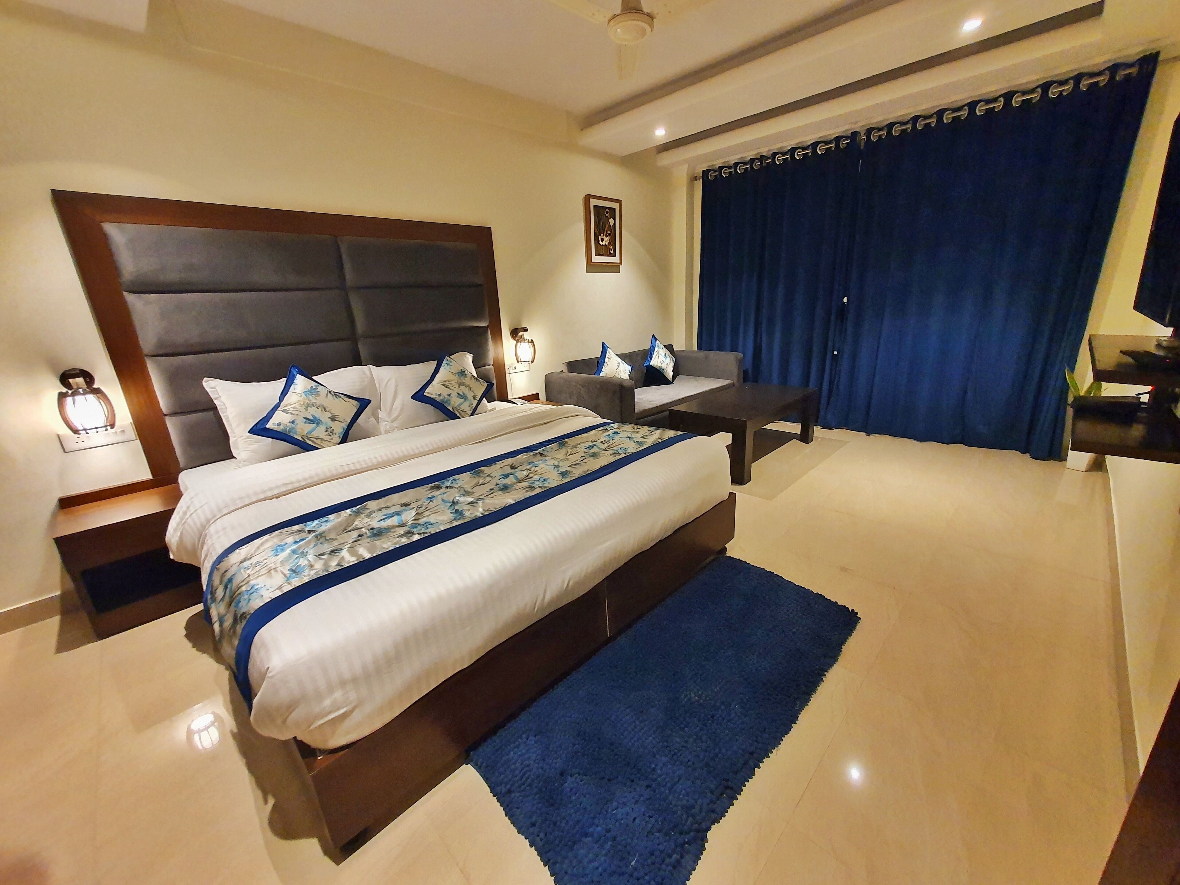 A hotel room with a large bed, a desk, a chair, a nightstand, a lamp, and a window with curtains.