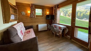 A cozy room with a sofa, a table with chairs, a TV, and large windows offering a view of a green landscape.