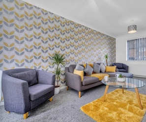 A modern living room with a patterned wallpaper, a grey sofa, a grey armchair, a glass coffee table, and a yellow rug.