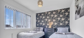 A bedroom with two beds, a dresser, a window with blinds, and a patterned wallpaper.