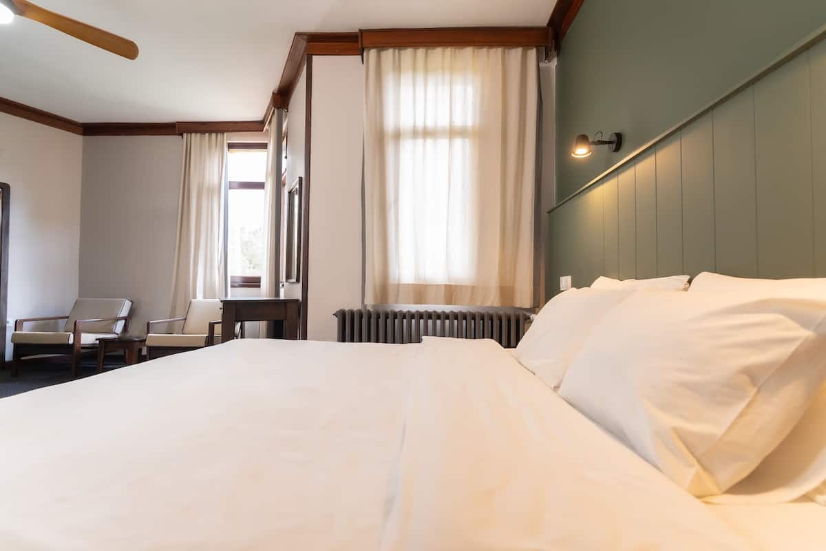 A hotel room with a large bed, two chairs, a desk, a radiator, and a window with curtains.