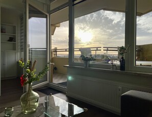 Ein Zimmer mit großem Fenster und Balkonblick, einem Glastisch, einer Vase mit Blumen und einer Heizung.