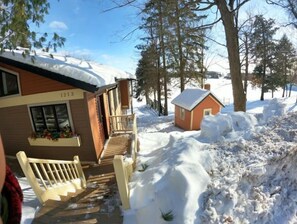 A wooden cabin with a balcony, a small attached shed, and a snow-covered path leading to the entrance.