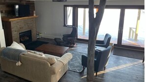 Ett vardagsrum med en öppen spis, en soffa, ett soffbord och ett stort fönster med utsikt över en veranda.