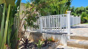 A stone pathway leading to a white picket fence with a garden area featuring tropical plants and a clear blue sky. - Bella Villa large apart for 4 with pool, jacuzzi @ 5 min walk to beach! (Samaná)