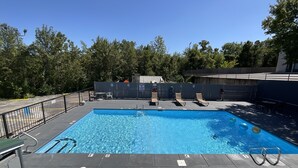 Une piscine extérieure rectangulaire à l’eau d’un bleu limpide, entourée d’une clôture noire. Des chaises longues et une table avec des chaises sont disposées le long du bord de la piscine.