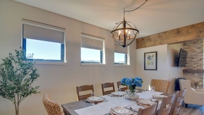Ein Esszimmer mit einem Holztisch, gedeckt für sechs Personen, umgeben von Holzstühlen. Zwei Fenster mit Jalousien, eine Kristallleuchter und ein kleiner Topfpflanze sind ebenfalls vorhanden.