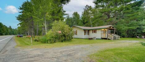 A small cabin with a porch and a staircase, surrounded by trees and a gravel driveway.