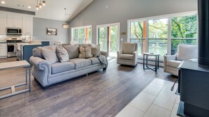 A spacious living area with a gray sofa and armchairs, a kitchen with white cabinets, and large windows offering a view of greenery. - Riverfront Salesville Cabin Rental w/ Shared Dock! (Mountain Home)