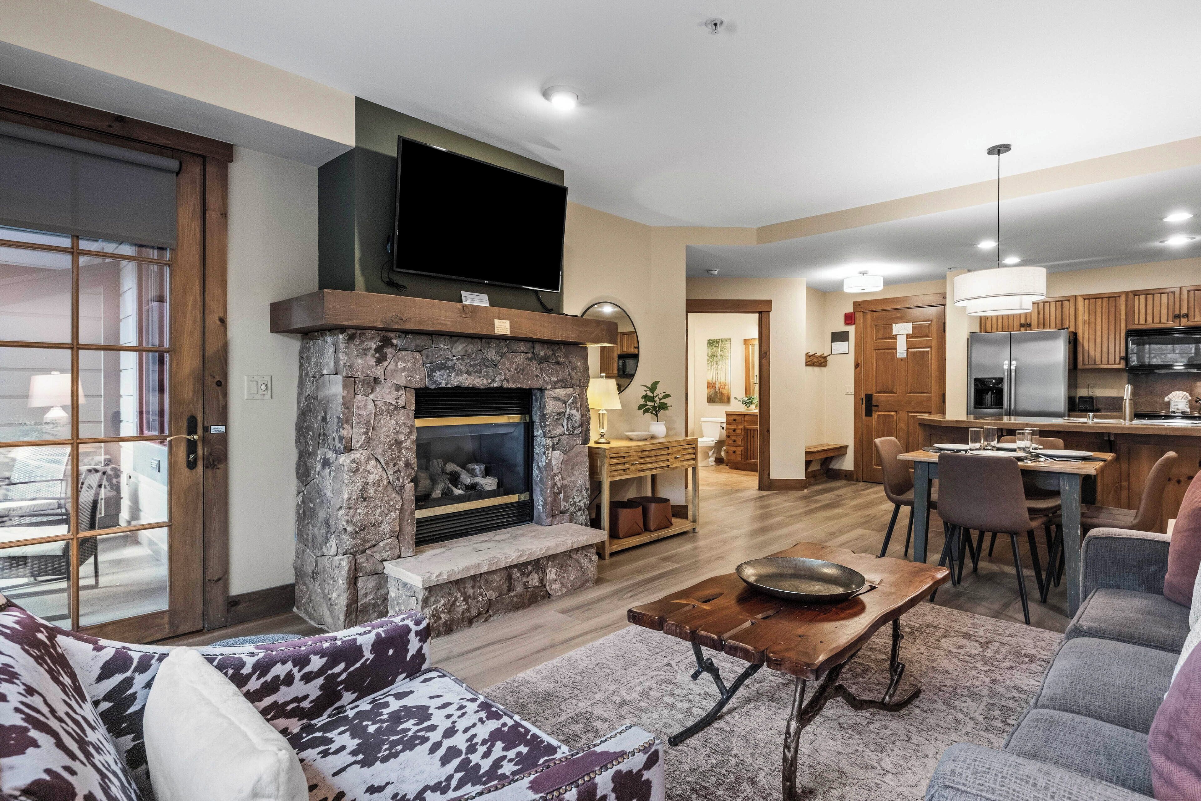 A living room with a stone fireplace, a television mounted above it, and a dining area with a kitchen in the background.