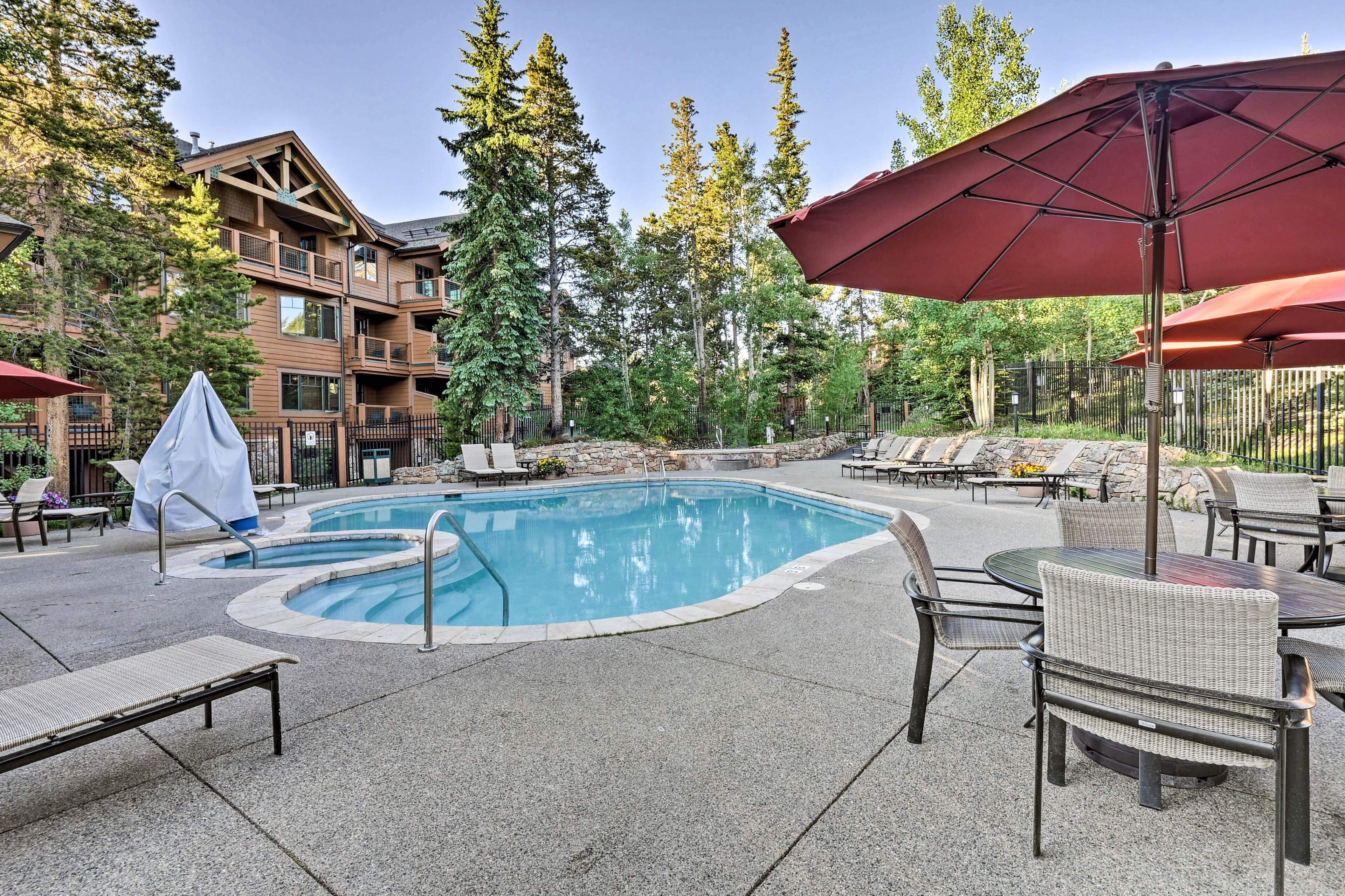 A pool area with lounge chairs and umbrellas, surrounded by tall trees and a building with balconies.