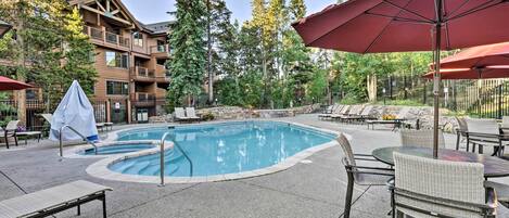 A pool area with lounge chairs and umbrellas, surrounded by tall trees and a building with balconies.
