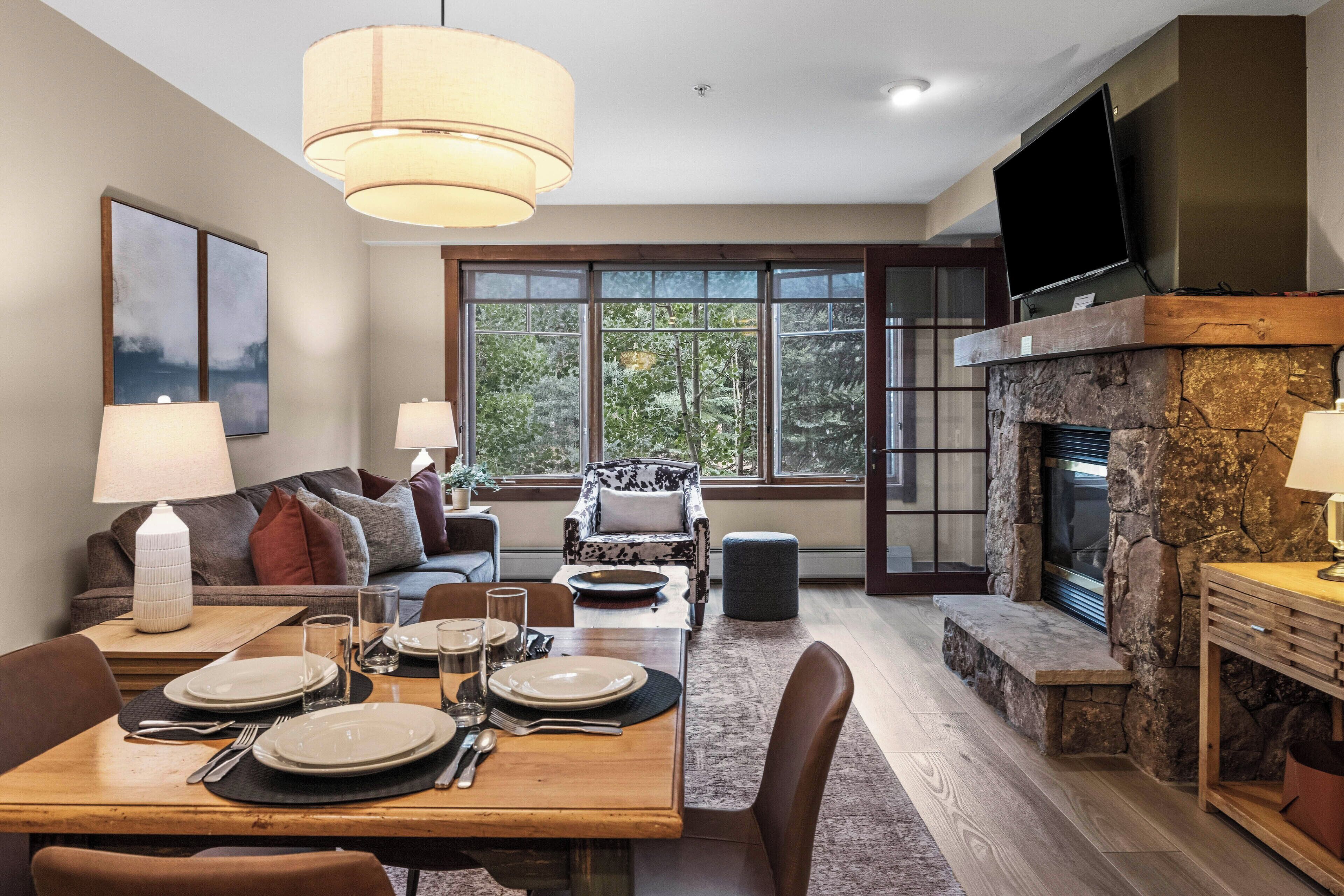 A spacious living room with a fireplace, a dining area with set tables, and large windows offering a view of trees.