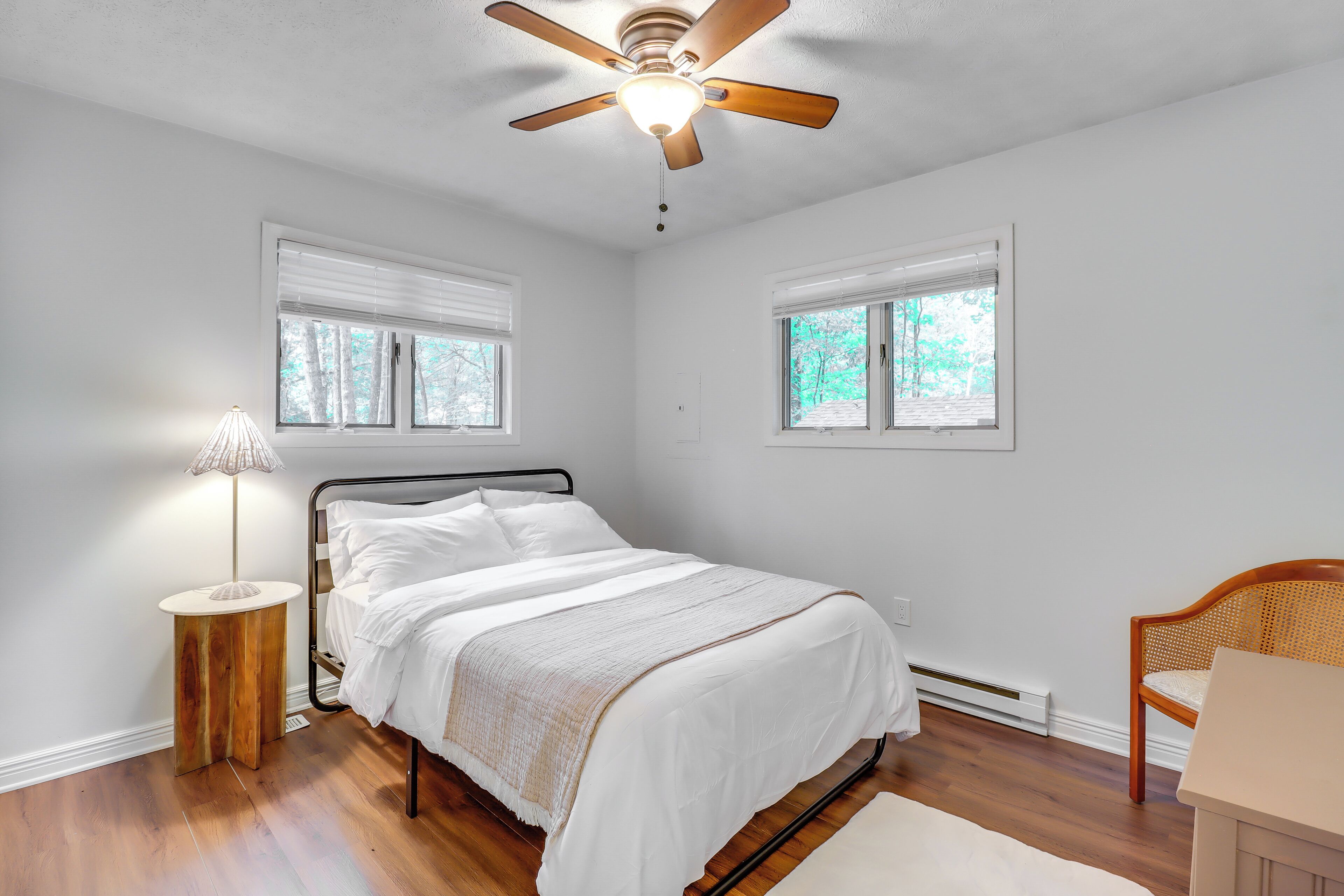 A bedroom with a bed, a ceiling fan, a bedside lamp, a chair, and two windows.