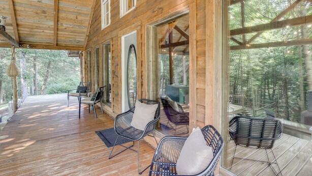 Une cabane en bois dotée d’un porche couvert, aménagée avec des chaises et une table en métal, et offrant une vue sur une forêt.